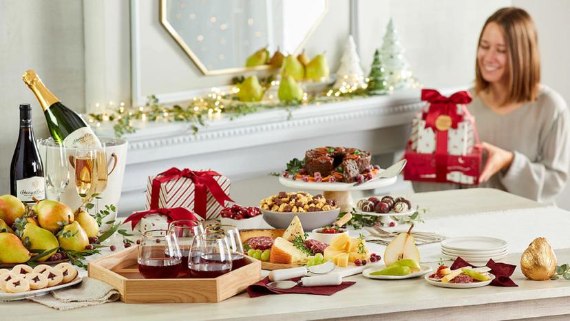 A photo of a fruitcake surrounded by other Christmas snacks and treats with a woman holding a stack of presents in the background