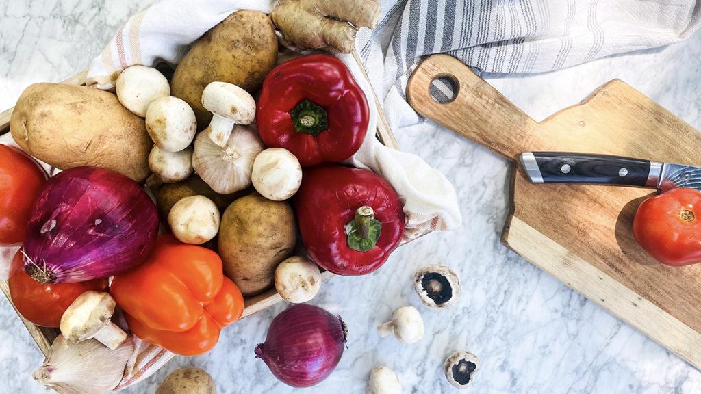 A photo of kebabs with a crate of seasonal vegetables next to a cutting board and knife